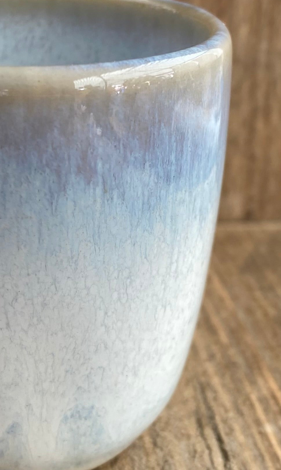 Close-up of a ceramic cup with a glossy glaze featuring a gradient from light blue to off-white, resting on a rustic wooden surface.